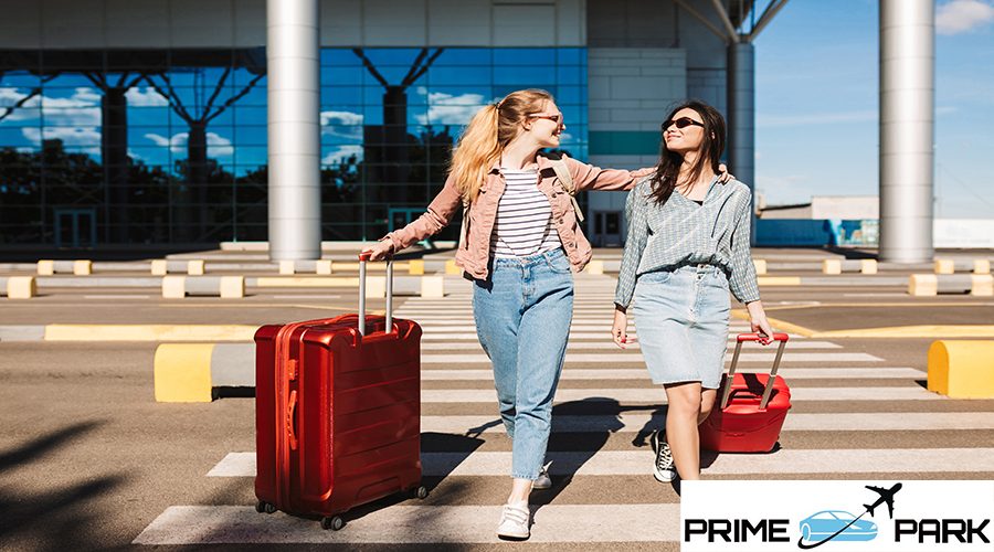 Beautiful stylish girls in sunglasses happily walking along pedestrian strip with red suitcases and airport on background