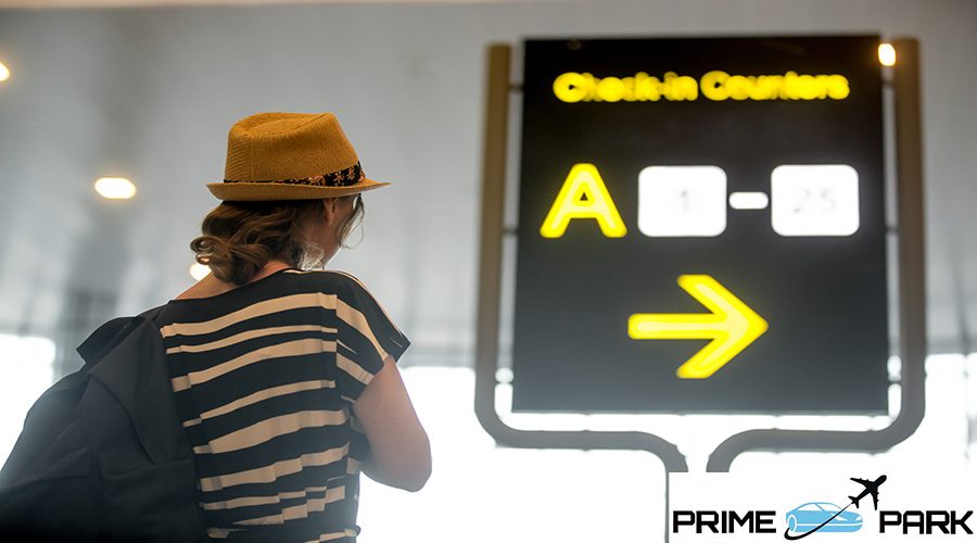 Young female traveller in straw hat with backpack in her 20s waiting for flight in modern airport terminal, looking at information sign for check in counters, back view