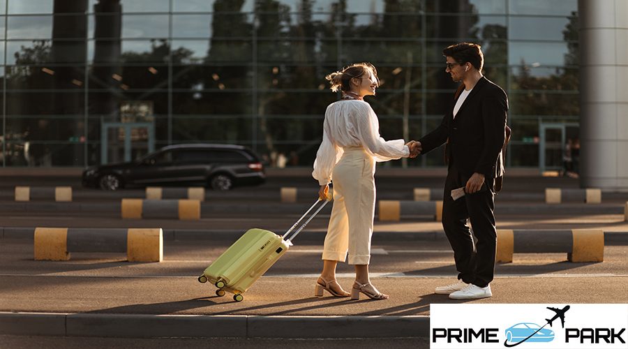 Happy blonde woman in white blouse, beige pants holds yellow suitcase and greets with man in black suit near airport.