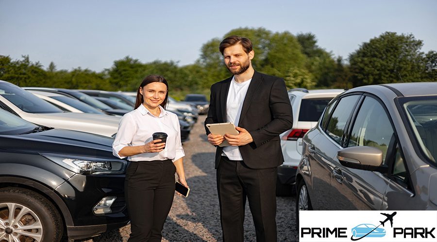 A male car dealer is showing a car to a woman, with a tablet in hand. They stand near a car lot on a sunny day.