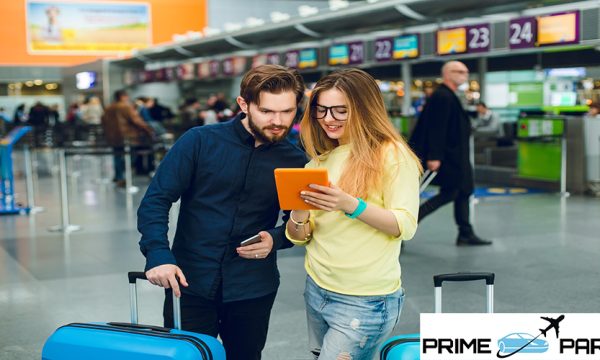 Young couple is standing between two suitcases in airport. She has long hair, glasses, sweater, jeans. He wears beard, black shirt with pants. They are reading on tablet.